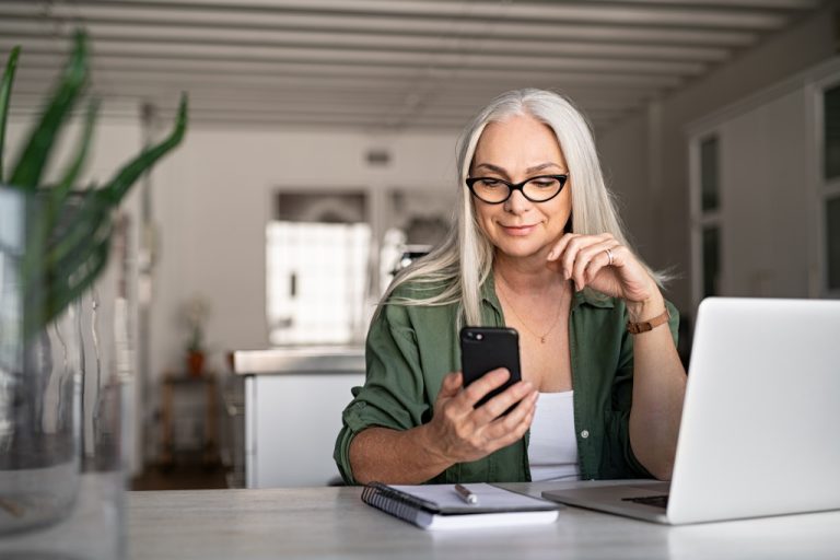 Happy senior woman using mobile phone while working at home with laptop. Smiling cool old woman wearing eyeglasses messaging with smartphone. Beautiful stylish elderly lady browsing site on cellphone.